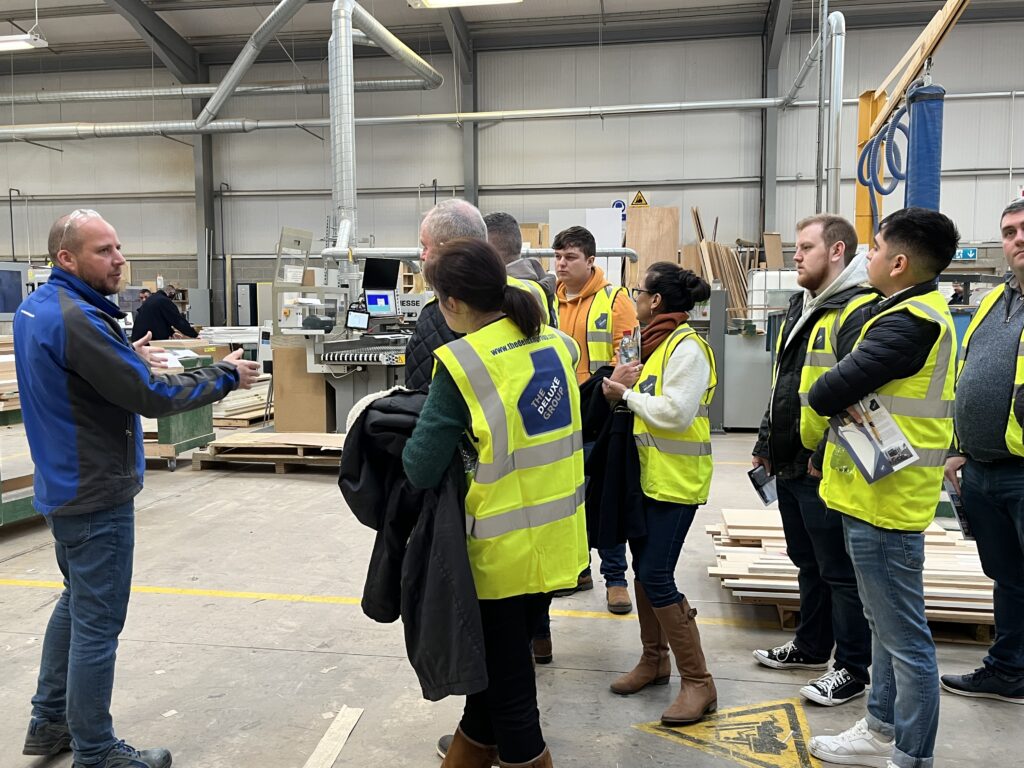A group of students in high vis jackets speaking with a member of staff from the Deluxe group on the factory floor. Computers and machinery can be seen in the background. 