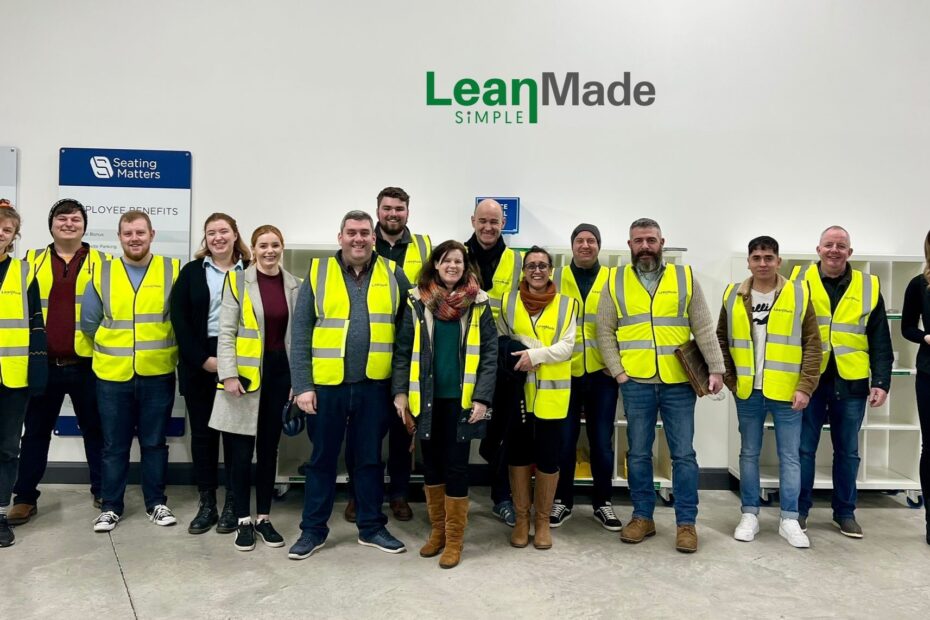 A group of staff and students line up against a white wall that has the words lean made simple on it.