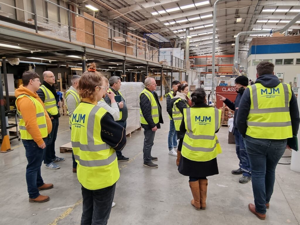 A group of students in high vis jackets stand in a semi circle on a factory floor listening to a talk from a member of staff at MJM Marine. 