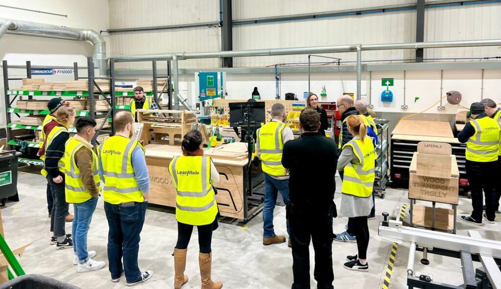 A group of students in high vis jackets stand in front of wood and work and machinery in Seating Matters. There is a member of staff talking to them. 